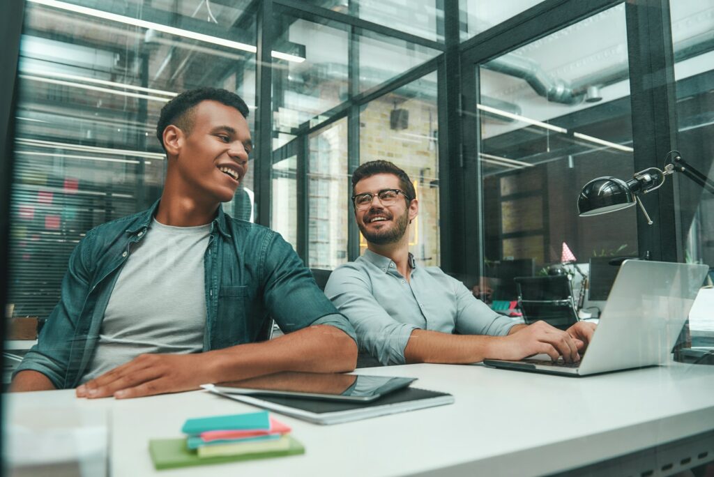 Positive atmosphere. Two young and handsome men talking with each other and smiling while working in